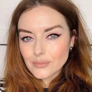 Op de afbeelding: Een close-up van een gezicht met rode eyeliner en lippenstift. De persoon heeft een lichte huid, groene ogen en rood haar. De eyeliner is in een gevleugelde vorm aangebracht. De lippen zijn nude.