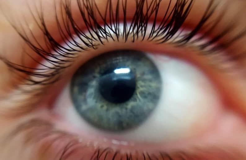 May include: Close-up of a human eye with a blue-green iris. The eye is surrounded by long, dark eyelashes. The image is well-lit, highlighting the details of the eye's texture and the surrounding skin.