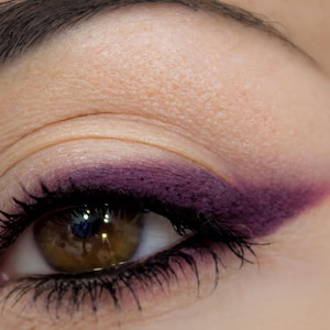 May include: A human eye featuring a bold, matte deep purple eyeshadow applied in a sharp, winged eyeliner shape along the upper lash line. The eyelid has a neutral beige base, and the dark brown iris is visible with black mascara on the lashes.