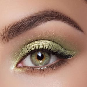 May include: A close-up of an eye featuring shimmering olive green eyeshadow applied across the eyelid, blended with a soft gold highlight at the inner corner. The look includes a dark green winged eyeliner, defined eyelashes, and a groomed eyebrow.