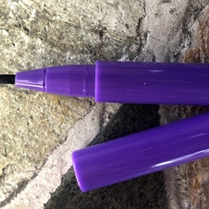 May include: A purple pen with a black felt tip and a matching cap. The cap is detached, revealing the pen's tip. The pen is lying on a textured, stone-like surface.