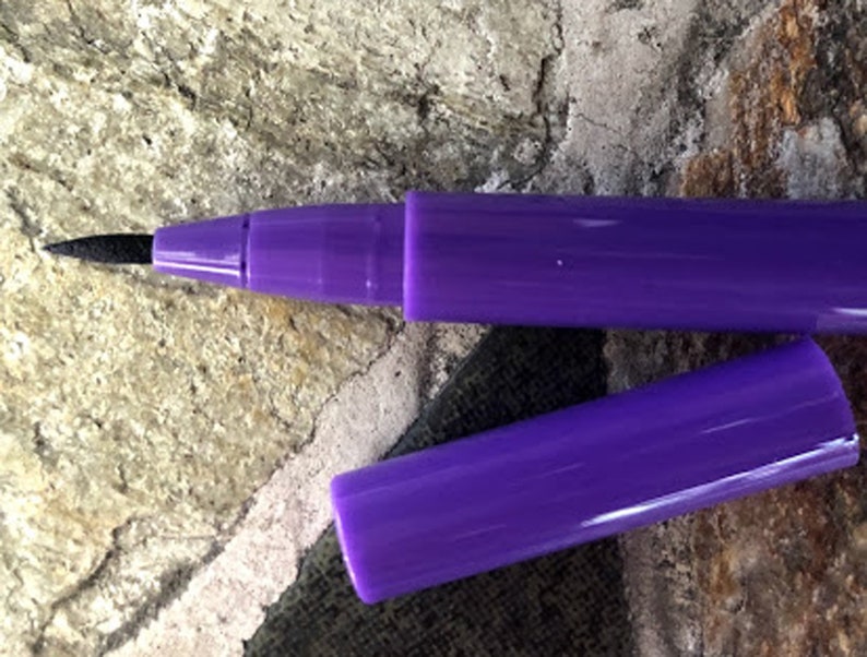 May include: A purple pen with a black felt tip and a matching cap. The cap is detached, revealing the pen's tip. The pen is lying on a textured, stone-like surface.
