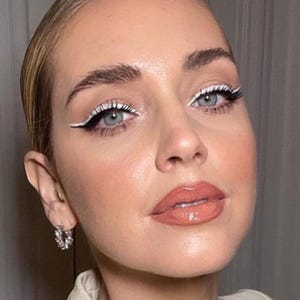 A close-up portrait of a woman with striking blue eyes, bold white eyeliner, and a bright red lipstick. She is wearing a beige top and has a pair of hoop earrings.