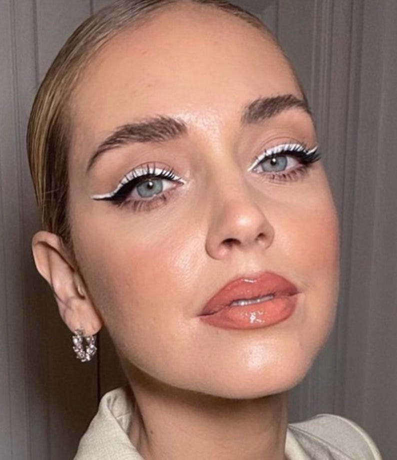 A close-up portrait of a woman with striking blue eyes, bold white eyeliner, and a bright red lipstick. She is wearing a beige top and has a pair of hoop earrings.