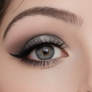 May include: Close-up of an eye with dramatic makeup. The eye features a smoky eye effect with shades of grey and black eyeshadow, a sharp black eyeliner wing, and long, dark eyelashes. The eyebrow is neatly groomed and filled in. The eye colour is a light blue-green.