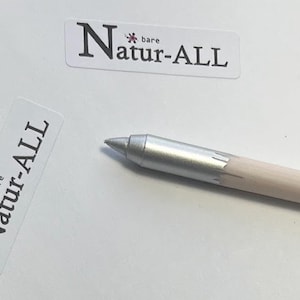 May include: A silver-tipped pencil with a natural wood barrel. The pencil is positioned on a white surface near two white rectangular stickers with the text "Natur-ALL". The word "bare" is above the word "Natur-ALL".