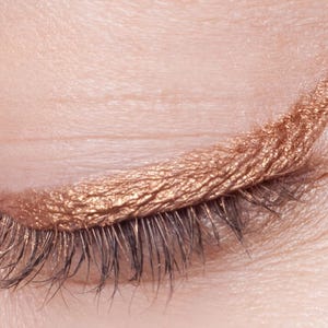Op de afbeelding: Een gesloten ooglid met een metallic koperkleurige vloeibare eyeliner aangebracht in een scherpe, gevleugelde lijn langs de bovenste wimperrand. De eyeliner heeft een getextureerde, glinsterende afwerking en de donkere wimpers zijn zichtbaar.
