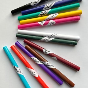 May include: An assortment of colorful eyeliner pencils in various shades, including black, blue, green, yellow, pink, white, and brown. Each pencil has a label with the text "Natur-ALL". The pencils are arranged on a white surface.