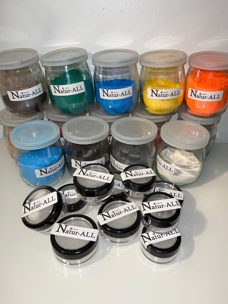 A collection of glass jars filled with various colored sand, labeled "Nature-ALL", arranged on a white surface.