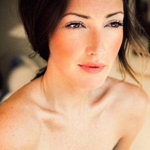 Può includere: Un ritratto in primo piano di una persona con la pelle chiara, i capelli castani e gli occhi verdi. La persona ha un trucco naturale con il fard sulle guance e un colore labbra rosa chiaro. L'illuminazione è morbida e calda.