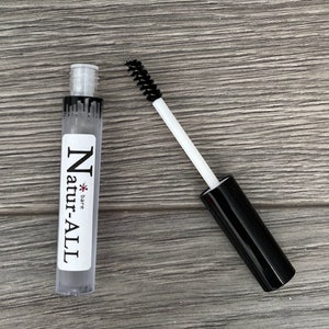 May include: A clear tube of Natur-ALL eyebrow gel with a black mascara wand. The tube has a white label with black text. The mascara wand has a black brush and a white handle. The background is a wooden surface.