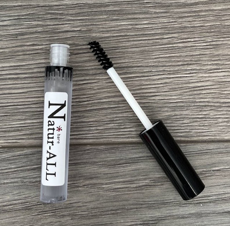 May include: A clear tube of Natur-ALL eyebrow gel with a black mascara wand. The tube has a white label with black text. The mascara wand has a black brush and a white handle. The background is a wooden surface.