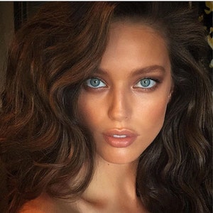Könnte beinhalten: Nahaufnahme eines Gesichts mit heller Haut, blauen Augen und braunem, welligem Haar. Die Person hat einen neutralen Gesichtsausdruck mit dezentem Make-up, einschließlich Lidschatten und Lippenfarbe. Die Beleuchtung ist weich.