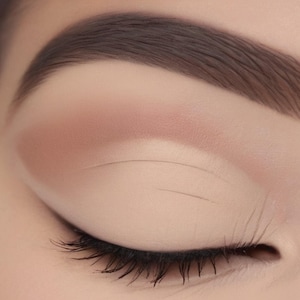 May include: A closed eyelid featuring a matte beige eyeshadow base with a soft brown transition shade blended into the crease. A thin, precise black eyeliner line is applied along the upper lash line, complemented by dark, defined eyelashes.