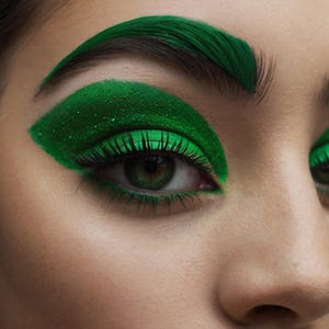May include: Close-up of an eye with vivid green eyeshadow and matching eyebrows. The eye has long, black eyelashes and a green iris. The makeup has a glittery finish, creating a striking and artistic look.