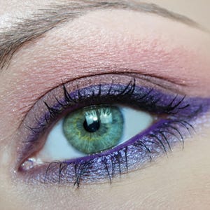 May include: A green iris eye featuring a makeup application with a soft pink eyeshadow on the upper lid, a shimmering purple eyeshadow on the lower lid, and a sharp purple winged eyeliner extending from the outer corner of the eye.