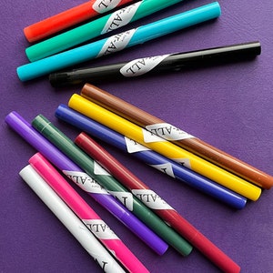 May include: A collection of colorful markers in various shades, including red, green, blue, black, brown, yellow, purple, and pink. Each marker has a white label with the word "ALL" printed on it. The markers are arranged on a purple surface.