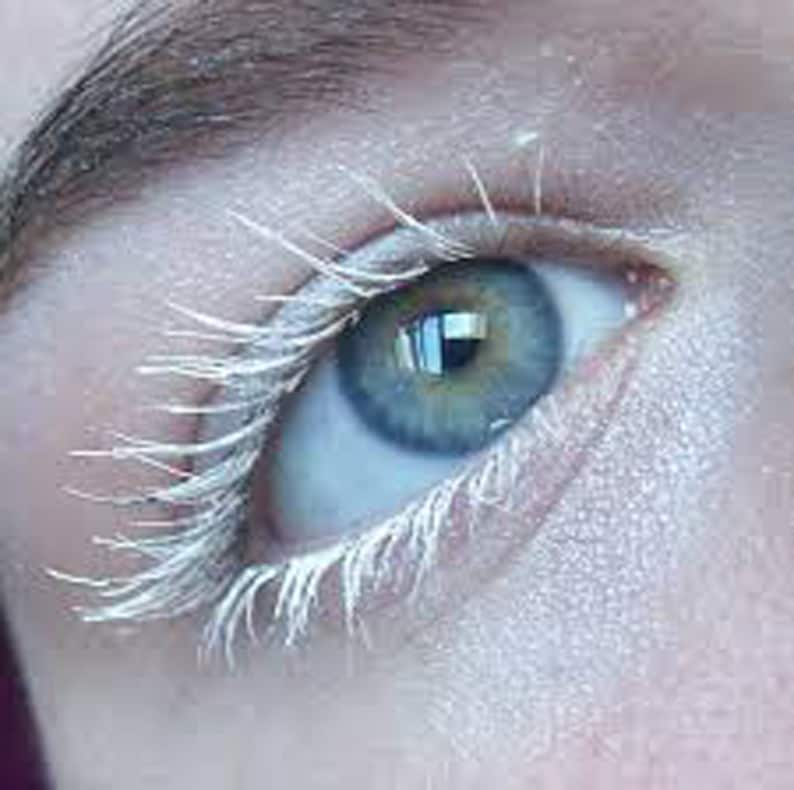 May include: Close-up of a human eye with blue-green iris and white eyelashes. The skin around the eye is pale. The eyelashes are coated in white mascara, creating a striking contrast with the eye color.
