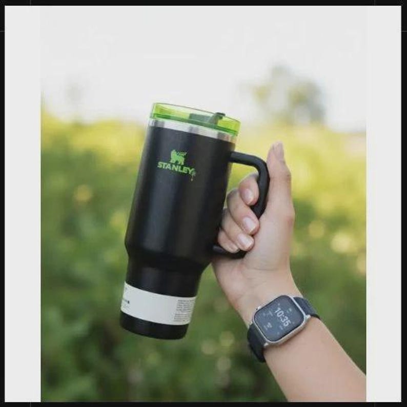 May include: Black Stanley insulated mug with a green lid and handle. The mug has the Stanley logo in green. The mug is held by a person wearing a smartwatch.