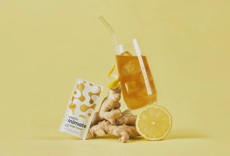 May include: A glass of iced drink with a straw, a packet of Unicity Unimate, ginger root, and a lemon slice, on a yellow background. The Unimate packet has a white and gold design. The ginger and lemon suggest a health drink.