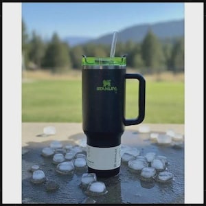 May include: A black Stanley travel mug with a green lid and handle, and a clear straw. The Stanley logo is in green. The mug is surrounded by ice cubes on a wet surface, with a blurred outdoor background.