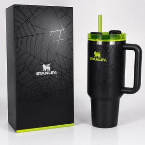 May include: Black Stanley travel mug with a green lid and straw, featuring a spiderweb design. The mug has a handle and the Stanley logo in green. A matching black box with a spiderweb design and the Stanley logo is next to the mug.