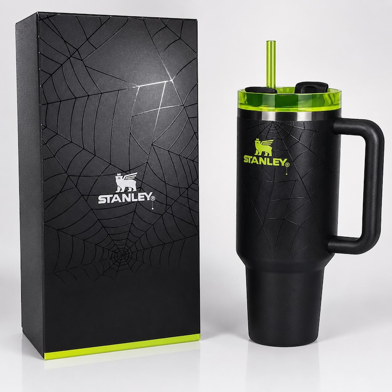 May include: Black Stanley travel mug with a green lid and straw, featuring a spiderweb design. The mug has a handle and the Stanley logo in green. A matching black box with a spiderweb design and the Stanley logo is next to the mug.