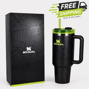 May include: Black Stanley tumbler with a green lid and straw, featuring a spiderweb design. The tumbler has a handle and the Stanley logo. A matching black box with a spiderweb design and the Stanley logo is next to the tumbler.