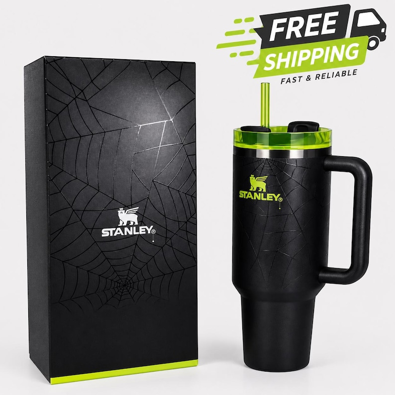 May include: Black Stanley tumbler with a green lid and straw, featuring a spiderweb design. The tumbler has a handle and the Stanley logo. A matching black box with a spiderweb design and the Stanley logo is next to the tumbler.