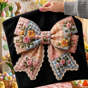 May include: A black fabric item featuring a large, decorative bow. The bow is composed of patchwork sections in pastel colors, including pink, blue, and peach gingham, and white polka dots. It is embellished with embroidered flowers and scalloped lace trim.
