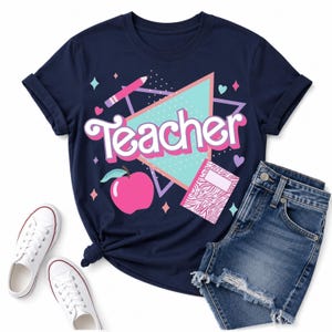 May include: A navy blue t-shirt featuring the word Teacher in white and pink script. The graphic includes a pink apple, a pencil, a notebook with a pink zebra print, and a light blue triangle. The shirt is paired with white sneakers and denim shorts.