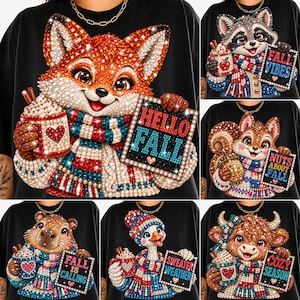 May include: A black t-shirt featuring a diamond art graphic of a fox wearing a scarf and holding a mug with a heart, next to a sign that says HELLO FALL. The image also displays other t-shirts with animals like a raccoon, squirrel, capybara, goose, and cow.