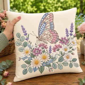 May include: A square cream-colored throw pillow featuring an embroidered design of a large butterfly with blue and pink patterned wings. The butterfly is surrounded by a detailed arrangement of lavender, white daisies, and green eucalyptus leaves.