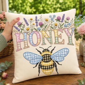 Peut inclure: Un coussin carré blanc cassé avec le mot HONEY brodé en lettres vichy multicolores. Au-dessus du texte se trouve un arrangement floral de marguerites, de lavande et de trèfle. Sous le texte, une grosse abeille aux ailes vichy.