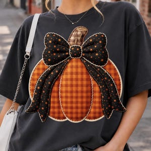 Może przedstawiać: Ciemnoszary t-shirt z dużą grafiką pomarańczowo-brązowej dyni w kratę. Dynia ozdobiona jest wyraźną czarną kokardą z małymi pomarańczowymi gwiazdkami. Wzór posiada efekt przeszywanego brzegu wokół konturu dyni na koszulce.
