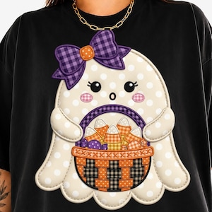 May include: A black t-shirt featuring a graphic of a cream-coloured ghost with white polka dots. The ghost wears a purple plaid bow and holds an orange and black woven basket filled with orange, yellow, and white candy corn and purple sweets.