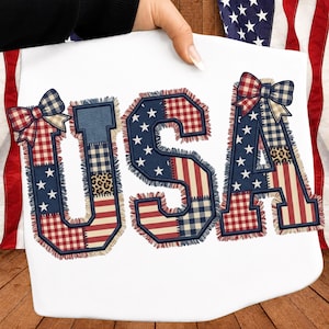 May include: A white t-shirt featuring the letters USA in a patchwork design. The letters are composed of red, white, and blue fabric patterns including stars, stripes, gingham, and leopard print, with frayed edges and a plaid bow on the U and A.