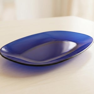 Puede incluir: Una bandeja de servir ovalada y lisa fabricada en vidrio azul cobalto intenso. El plato presenta un centro cóncavo poco profundo con un borde redondeado ligeramente elevado, mostrando un acabado translúcido que capta la luz en su superficie.