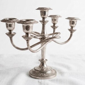 May include: A silver-toned metal candelabra featuring five candle holders arranged in a tiered design. The central pillar is elevated, surrounded by four curved arms. The base and the rims of the candle cups feature intricate embossed floral detailing.