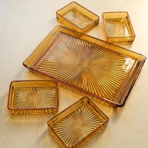 Puede incluir: Un conjunto de cinco bandejas de vidrio de color ámbar con un patrón de rayos de sol que irradia desde el centro. La colección incluye una bandeja rectangular grande y cuatro bandejas rectangulares más pequeñas, todas con bordes elevados.