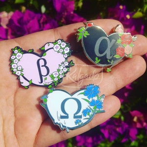 May include: Three heart-shaped enamel pins. One is light pink with a white floral border and a beta symbol. Another is black with a silver alpha symbol and red roses. The third is white with a blue omega symbol and blue flowers.