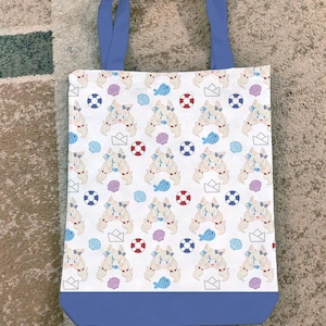 Puede incluir: Bolso tote blanco con asas azules y base azul. El bolso presenta un patrón repetido de un personaje de dibujos animados, conchas marinas, peces, salvavidas y barcos de papel. El bolso está sobre una alfombra de color neutro.