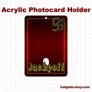 May include: Acrylic photocard holder with a dark red background and gold trim. The holder features the word "Jackpot!" in gold lettering at the bottom and a logo at the top. A small hole is at the top for hanging.