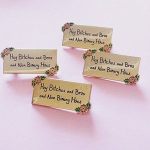 May include: Four gold-colored enamel pins with a rose design and the text "Hey Bitches and Bros and Non Binary Hoes" on a pink background.