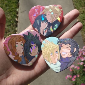 May include: Three heart-shaped pins featuring anime characters. The first pin is red with a couple embracing. The second pin is pink with a woman with long brown hair and a man with black hair. The third pin is blue with a man with blonde hair and a man with black hair.