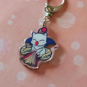 May include: A clear acrylic keychain featuring a cartoon character with a white body, red cheeks, and a red cherry on its head. The character has blue bat-like wings and is holding a broom. The words "SAVE" and "GAME" are on the keychain.