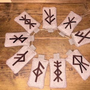 May include: A circular garland of nine rectangular felt pouches in a light beige color, each featuring a unique dark brown embroidered rune symbol. The pouches are connected by a thin, natural-colored twine string looped through small fabric tabs.