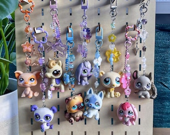 Puede incluir: Una colección de diez llaveros decorativos con figuras de animales en miniatura unidas a cadenas de cuentas coloridas con varios dijes que incluyen estrellas, flores y lazos, todos suspendidos de un tablero perforado de madera clara.