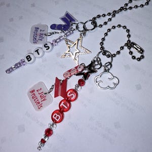 Pode incluir: Dois chaveiros de contas com correntes de bolas prateadas. Um chaveiro tem contas de letras brancas que soletram "BTS" e um pingente transparente com o texto "2 July Brussels". O outro chaveiro tem contas de letras vermelhas que soletram "BTS" e um pingente transparente com o texto "2 July Brussels".
