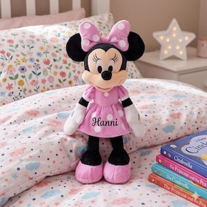 May include: A Minnie Mouse plush doll wearing a pink dress with white polka dots and a matching bow. The doll has the name "Hanni" embroidered on the dress. The doll is on a bed with a floral patterned comforter and pillows.